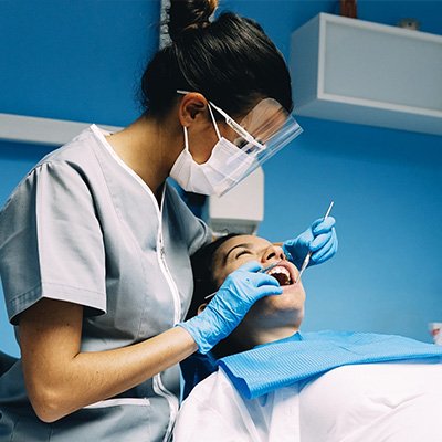 Dentist or hygienist wearing a face shield and mask, performing a dental examination.