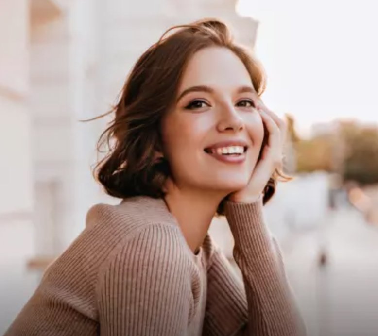 A happy young woman with short, wavy brown hair, smiling broadly and touching her cheek, with a blurred outdoor background.