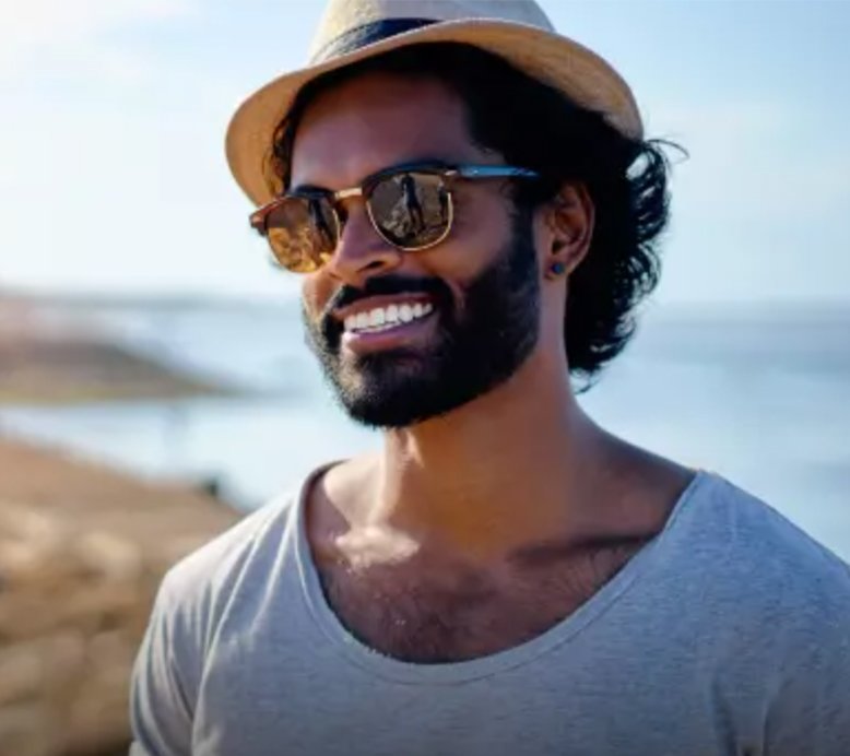 A man with dark, curly hair, a beard, and sunglasses, smiling broadly while looking slightly to the right, with a blurred outdoor background of water and land.