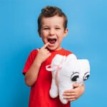 Happy young boy pointing to his tooth while holding a plush tooth toy, representing pediatric dentistry or fillings.