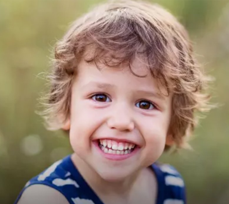A close-up of a young child with curly brown hair, wearing a striped shirt, smiling widely at the camera, with a blurred green background.