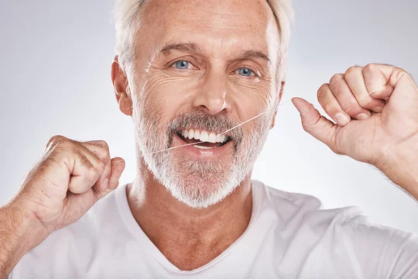 A smiling man with white hair and a beard, holding dental floss between his teeth, demonstrating good oral hygiene.