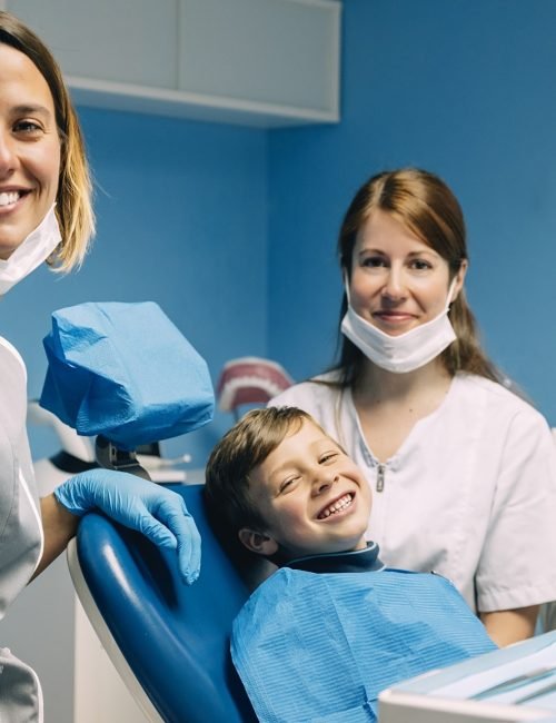 Happy young boy smiling in a dental chair with two friendly female dental professionals.